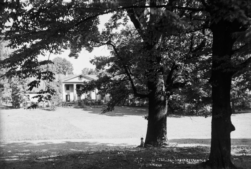 czarno biała fotografia budynku tzw. Ludwikówka. Po prawej chłopczyk siedzący pod drzewem  to Jerzy Święch, syn Michała Święcha autora fotografii