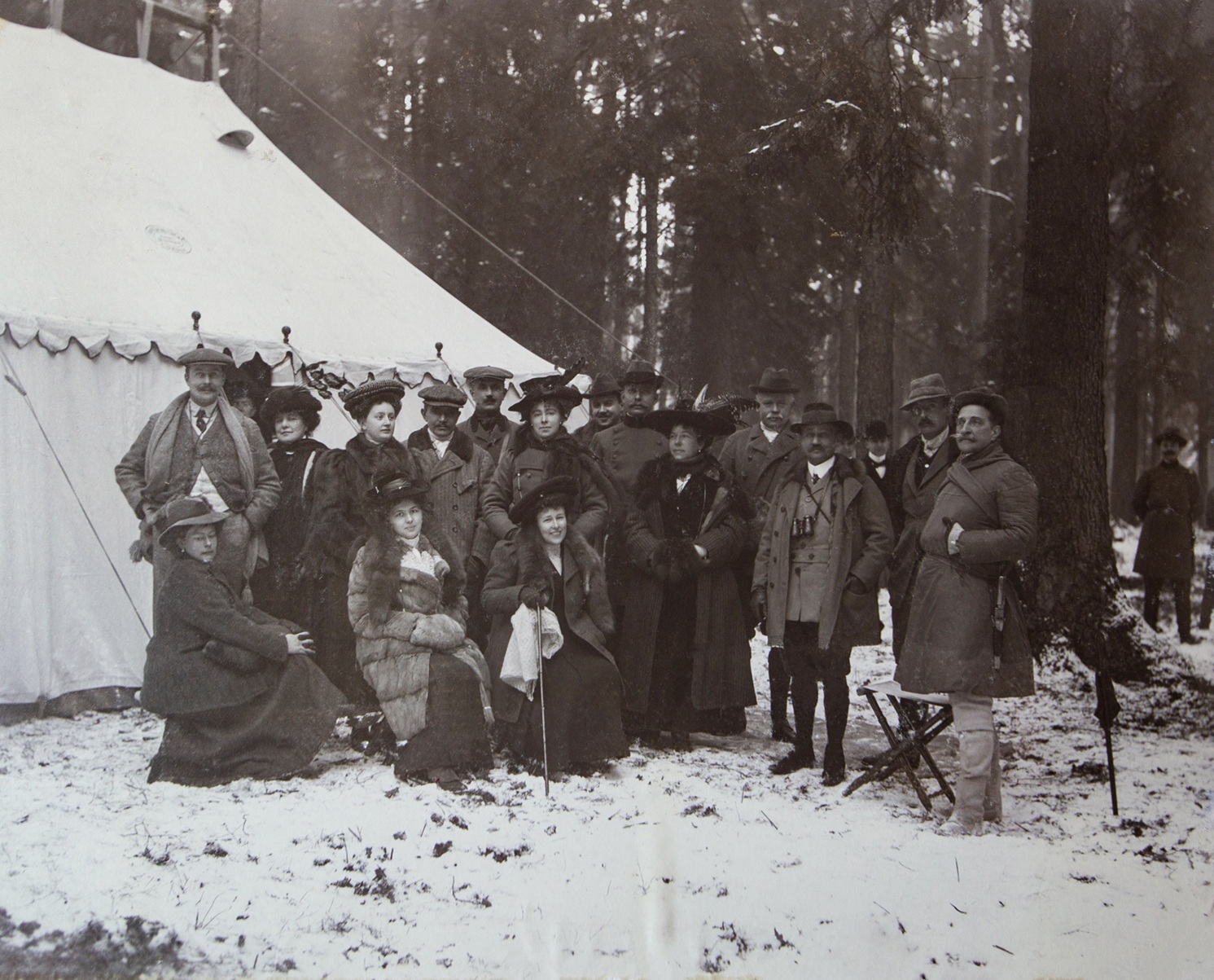 Zdjęcie przedstawia grupę elegancko ubranych mężczyzn i kobiet zgromadzonych na polowaniu w lasach pszczyńskich 13 grudnia 1908 roku. W tle widać duży namiot myśliwski oraz wysokie drzewa pokryte lekką warstwą śniegu. Część osób ma na sobie futra i kapelusze, a kilku mężczyzn nosi charakterystyczne stroje myśliwskie. Na pierwszym planie dwie kobiety siedzą na śniegu, trzymając w dłoniach laski i parasolkę. Wśród zgromadzonych księżna Daisy Hochberg von Pless. Atmosfera zdjęcia sugeruje towarzyskie spotkanie zorganizowane w zimowej scenerii.